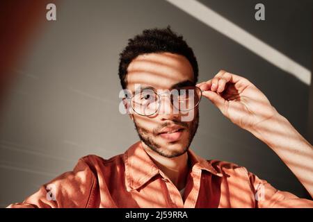 Selfie portrait of handsome young mixed race guy adjusting eyeglasses against isolated background, light through blinds effect Stock Photo