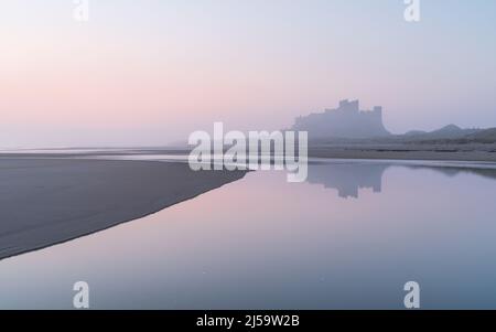 A beautiful pastel sunrise during a misty morning on Bamburgh Beach, with the famous castle reflected in a perfectly calm tidal pool. Stock Photo