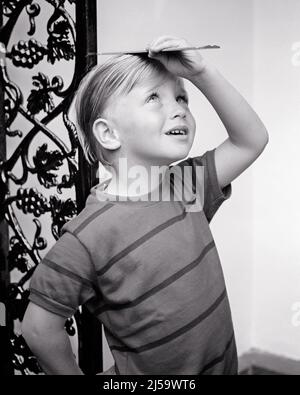 1960s YOUNG BLONDE BOY TRYING TO MEASURE HIS OWN HEIGHT WITH A RULER - j12487 HAR001 HARS INDEPENDENT STATURE COMEDY CURIOUS GROW T-SHIRT CHILDHOOD GROWTH JUVENILES LOOKING UP OWN ROOM DIVIDER TEE SHIRT BLACK AND WHITE CAUCASIAN ETHNICITY HAR001 OLD FASHIONED Stock Photo