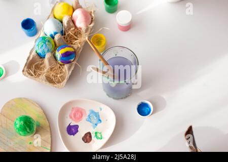 On a white table, in daylight, there are brushes in a glass of water, multi-colored eggs in a tray, jars of paint Stock Photo