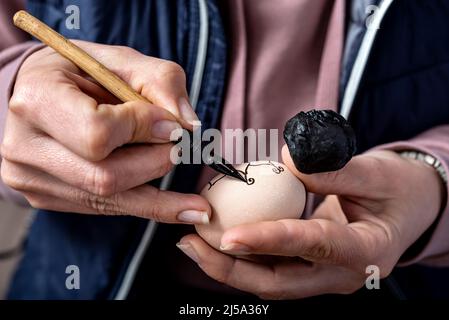 Female hands traditionally paint eggs with floral patterns using a fountain pen and wax for Easter. Easter preparation concept. Stock Photo