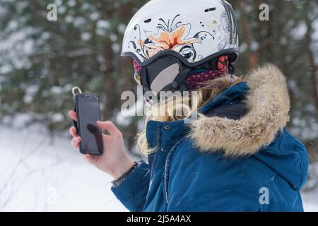 The girl is holding a mobile phone in her hands. She is wearing a ski helmet and standing on the side of a mountain. Stock Photo