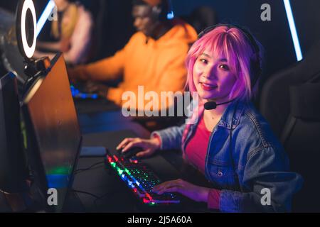 PC bang. Cute laid-back Asian pink-haired girl looking at camera while wearing professional headset, and playing on desktop computer. High quality photo Stock Photo