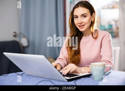 Portrait of young woman sitting at table in living room looking laptop screen Stock Photo
