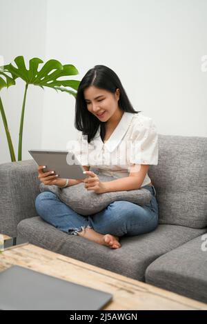 Attractive Asian woman spend her weekend at home, relaxes on sofa, enjoys using tablet, scrolling on online shopping application. Stock Photo