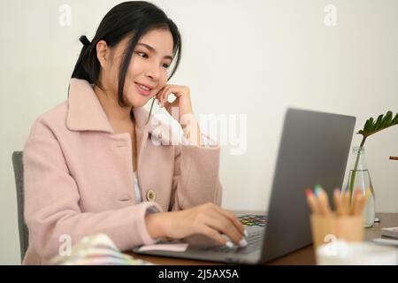 Attractive Asian female graphic designer working on laptop computer in minimalist office workspace. Stock Photo