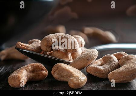 cashews on a spoon and on a wooden table with a black bowl and dark wooden wall in the background dark food or moody light macro photography Stock Photo