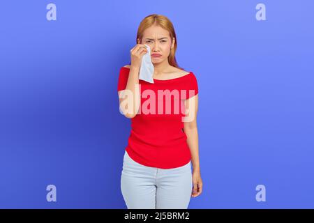 Portrait of sad woman she is crying wiping tears from eyes with tissue isolated on purple background Stock Photo