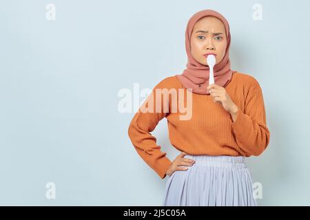 Beautiful Asian woman in brown sweater and hijab covering her mouth with spoon, wants to try delicious food isolated on white background Stock Photo