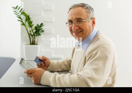 Handsome cheerful senior man using digital tablet sitting at the table in the living room. Leisure activities, spending time, happy retirement and Stock Photo