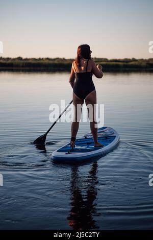 Woman of middle-age standing on paddle board having one oar in hands to steer board looking up on crystal blue lake wearing swimming dress. Active Stock Photo