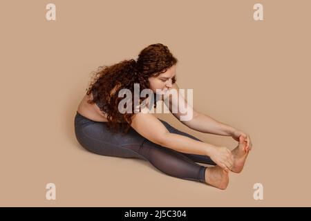 Red curly haired melancholy calm fat overweight woman flabs not hamstring muscles and spin, stretching. Gray sportswear Stock Photo