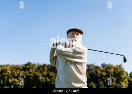 low angle view of bearded senior man in flat cap holding golf club while playing outdoors Stock Photo