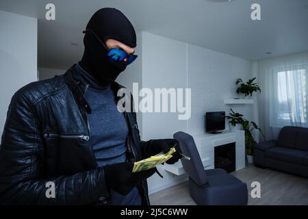 Robber man dressed in black hoodie stands with disguised face and holds a lot of money in his hands, stole a large amount, a thief man stole a TV Stock Photo