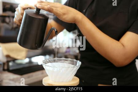 A female cafe operator wearing an apron pours hot water over roasted coffee grounds to prepare coffee for customers in the shop. Stock Photo