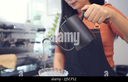 A female café operator wearing an apron pours hot water over roasted coffee grounds to prepare coffee for customers in the shop. Stock Photo