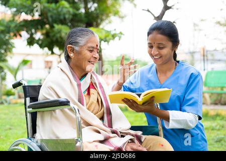 Nurse and senior woman on wheel chair laughing by reading book or novel at park - concept of relaxation, professional occupation, therapy or treatment Stock Photo