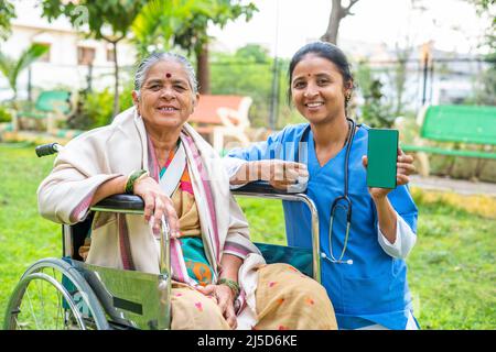 doctor showing green screen mobile phone wile patient sitting on wheelchair at park - concept of technology, healthcare app promotion and Stock Photo