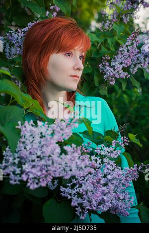 Young woman with bouquet of lilacs in spring. Red-haired lady sniffs purple flowers in nature. Portrait of attractive female in flower garden. Stock Photo