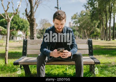 Surfing the internet, the young man is sitting on a bench in the spring weather, surfing the Internet with his phone, social media addiction and the c Stock Photo