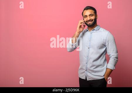 Indian man in jeans shirt talking phone, calling to somebody, looking at the camera with happy facial expression. Indoor studio shot isolated on pink background Stock Photo