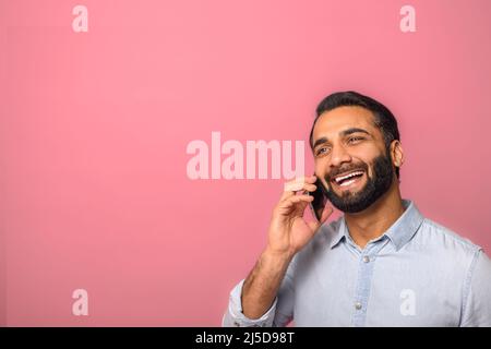 Cheerful optimistic Young Indian bearded man talking phone, calling to discuss plans, looking away with dreamy facial expression. Indoor studio shot isolated on pink background Stock Photo