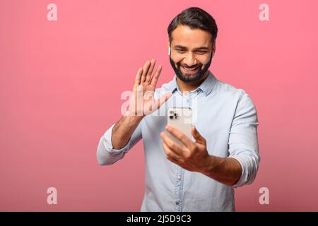 Young handsome smiling Indian man making video call on the smartphone, waving, chatting via cell phone or making selfie isolated on pink background Stock Photo