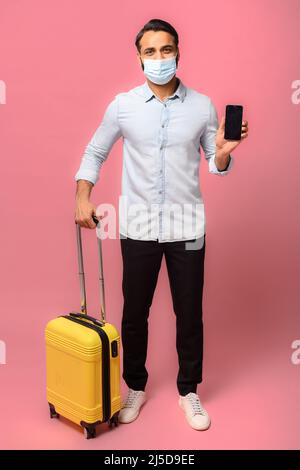 Full length Indian man in medical mask standing over pink background holding yellow handbag for travel and cell phone with empty screen, concept of contactless ticket display, safe passage of customs Stock Photo