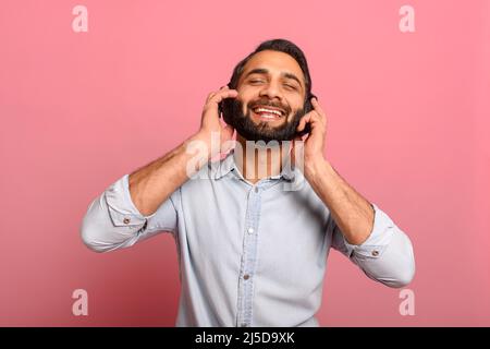 Studio portrait of relaxed cheerful Indian man wearing and holding wireless headphones isolated on pink, enjoying listening to favorite music soundtrack, wellbeing, meditation concept Stock Photo
