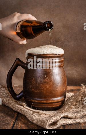 Beer clay brown mug with beer on a wooden table on a dark background. A stream of light beer pours into a beer glass Stock Photo