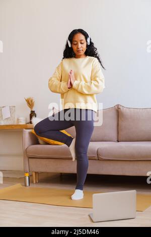 African-American female in headphones standing in tree pose yoga while following tutorial on laptop Stock Photo