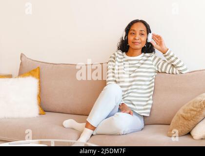 Smiling African-American female listening to music in headphones while sitting on couch Stock Photo