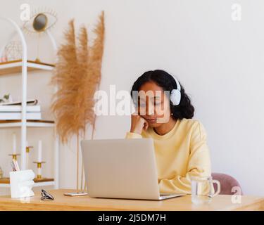Sleepy African-American female student in headphones attending online lecture on laptop at home Stock Photo