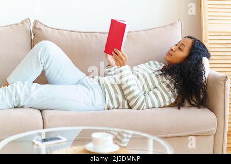 African-American lying down on sofa and reading book in red cover. Slow paced lifestyle Stock Photo