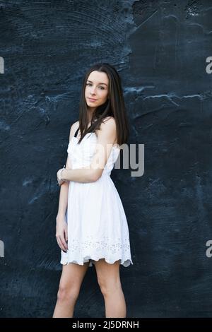 A beautiful teen brunette girl with a serious look leaning against a black wall Stock Photo