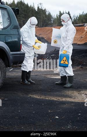 Two ecologists in protective workwear and rubber boots standing at car on polluted land and holding containers for toxic substances Stock Photo