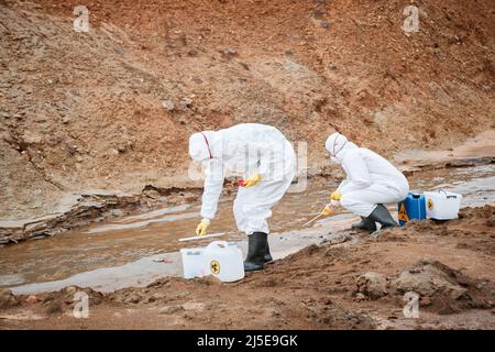 Two environmental scientists in protective workwear and masks taking samples of toxic water while examining it on polluted area Stock Photo