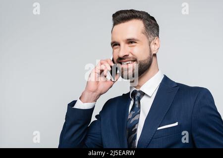 happy economist in blue blazer looking away while talking on smartphone isolated on grey Stock Photo