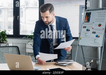 brunette economist holding papers and looking at laptop in office Stock Photo