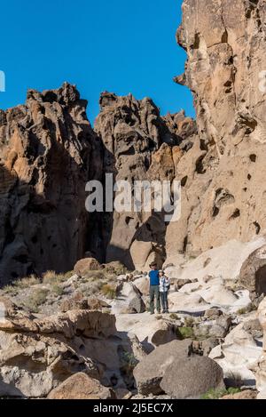 A father and son stand on boulders at Banshee Canyon with Hole in the Wall cliffs behind them, hiking Rings Loop trail in Mojave National Preserve. Stock Photo