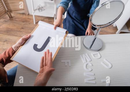 Cropped view of speech therapist and kid pointing at letter on clipboard near mirror in classroom Stock Photo