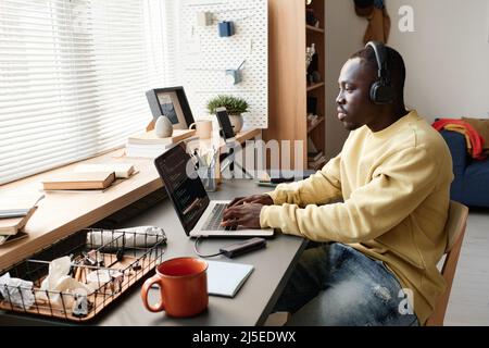 Concentrated young African-American coding specialist in headphones sitting at table against window and using laptop while creating app Stock Photo