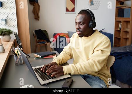 Serious young black man in headphones and yellow sweater sitting at table in students room and using laptop Stock Photo