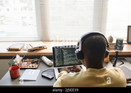 Rear view of black programmer in wireless headphones sitting at desk with hard drive and using laptop while working on java script Stock Photo