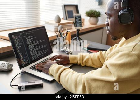Concentrated African American app developer in yellow sweater sitting at table and working on code using laptop Stock Photo