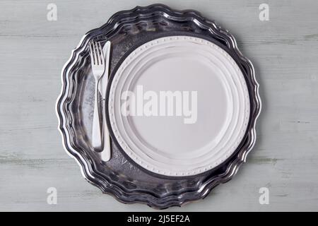 White plate with cutlery on a shiny metal tray on a light wooden table Stock Photo