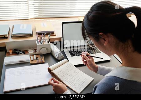 Over shoulder view of Asian student in glasses sitting at table and making notes in notepad while solving math problem Stock Photo