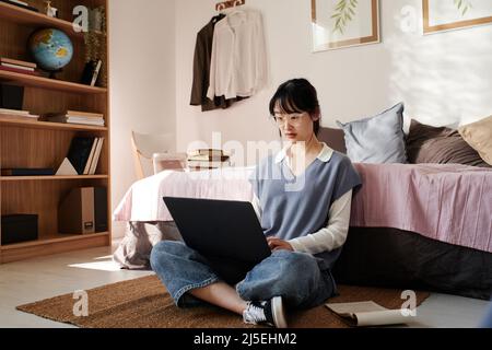 Serious young Asian female student in eyeglasses sitting with crossed legs on floor and reading article on portable computer Stock Photo