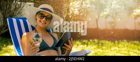 young attractive woman wearing bikini relaxing in sun lounger in garden. drinking wine and using digital tablet. copy space Stock Photo