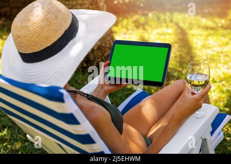 mockup of woman wearing bikini using digital tablet and drinking wine while relaxing in sun lounger in garden Stock Photo